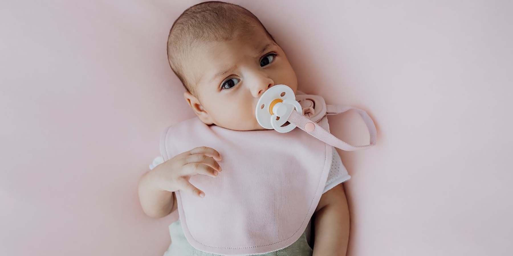A baby wearing a pink Gigi Bib. The baby has a dummy in his mouth, secured to the bib with the Gigi Accessory Tether