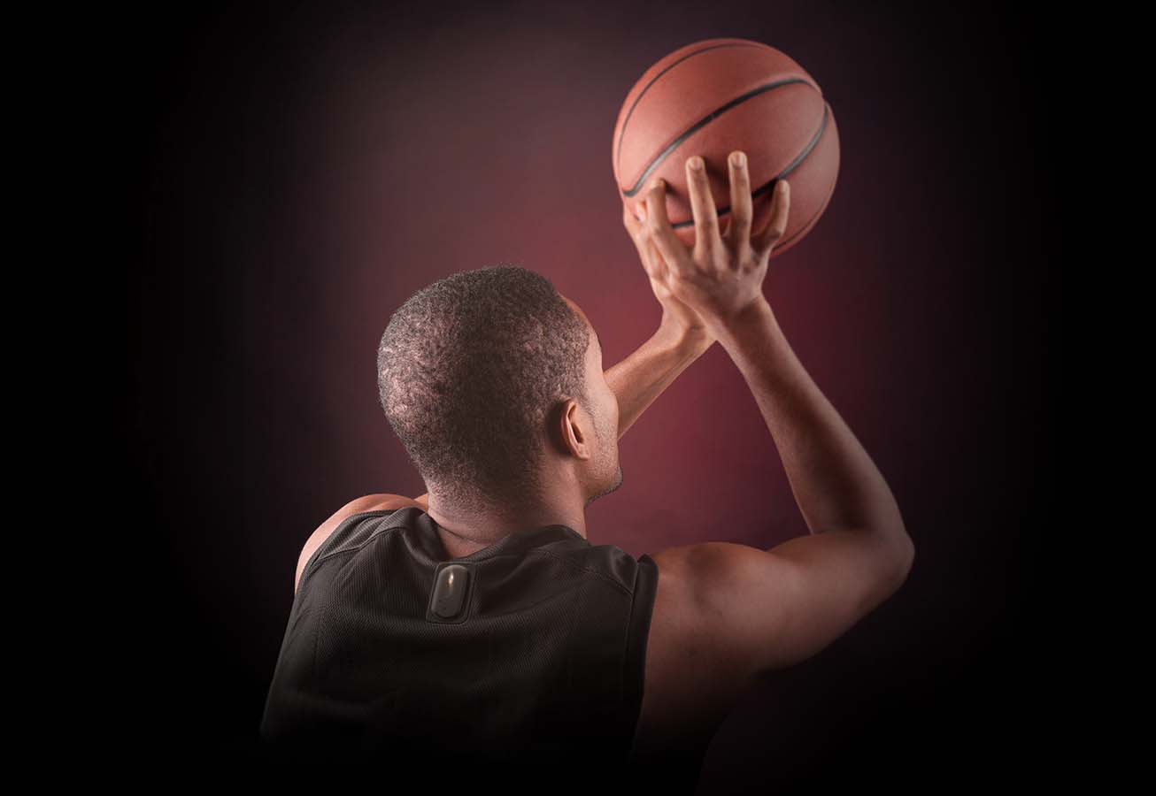 A photograph of a man laying up to shoot a basketball. The photo is taken from behind. The tracker is mounted to singlet.