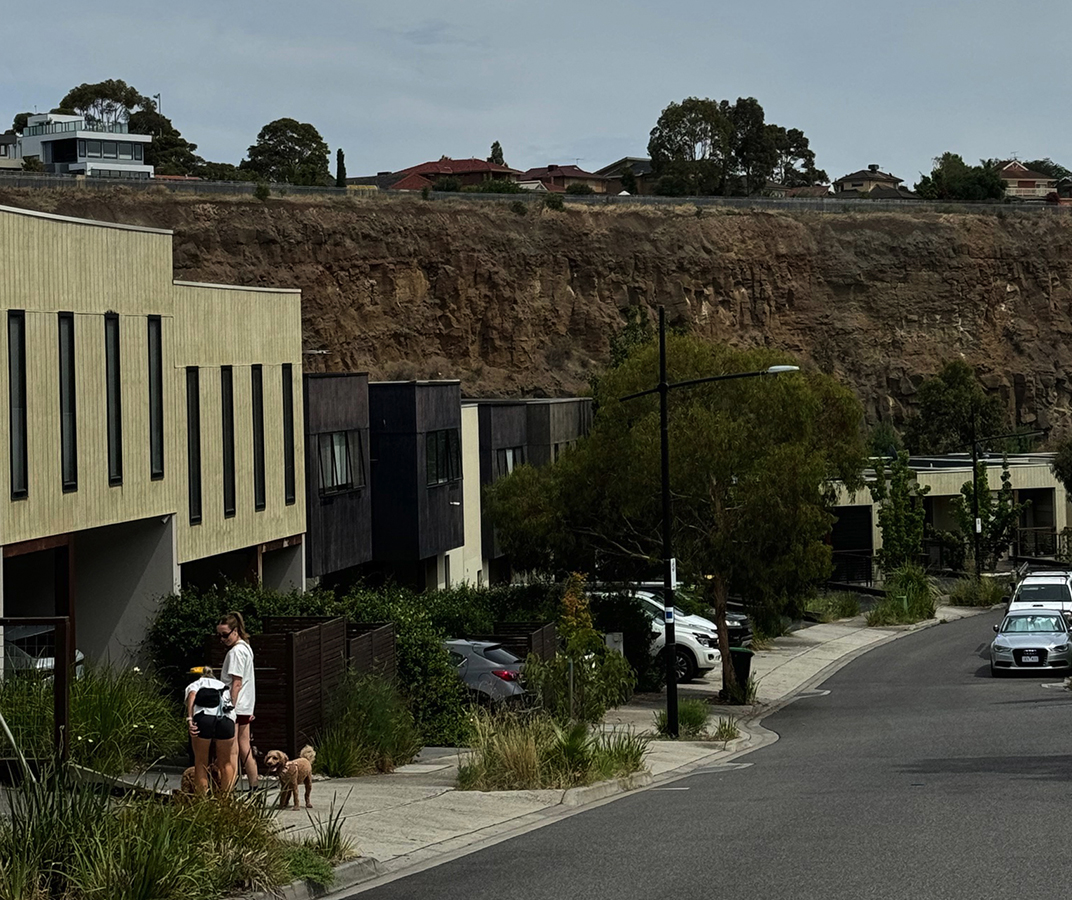 A row of timber terrace houses stepping down a slope with girls walking a dog and the quarry cliff face in the distance.