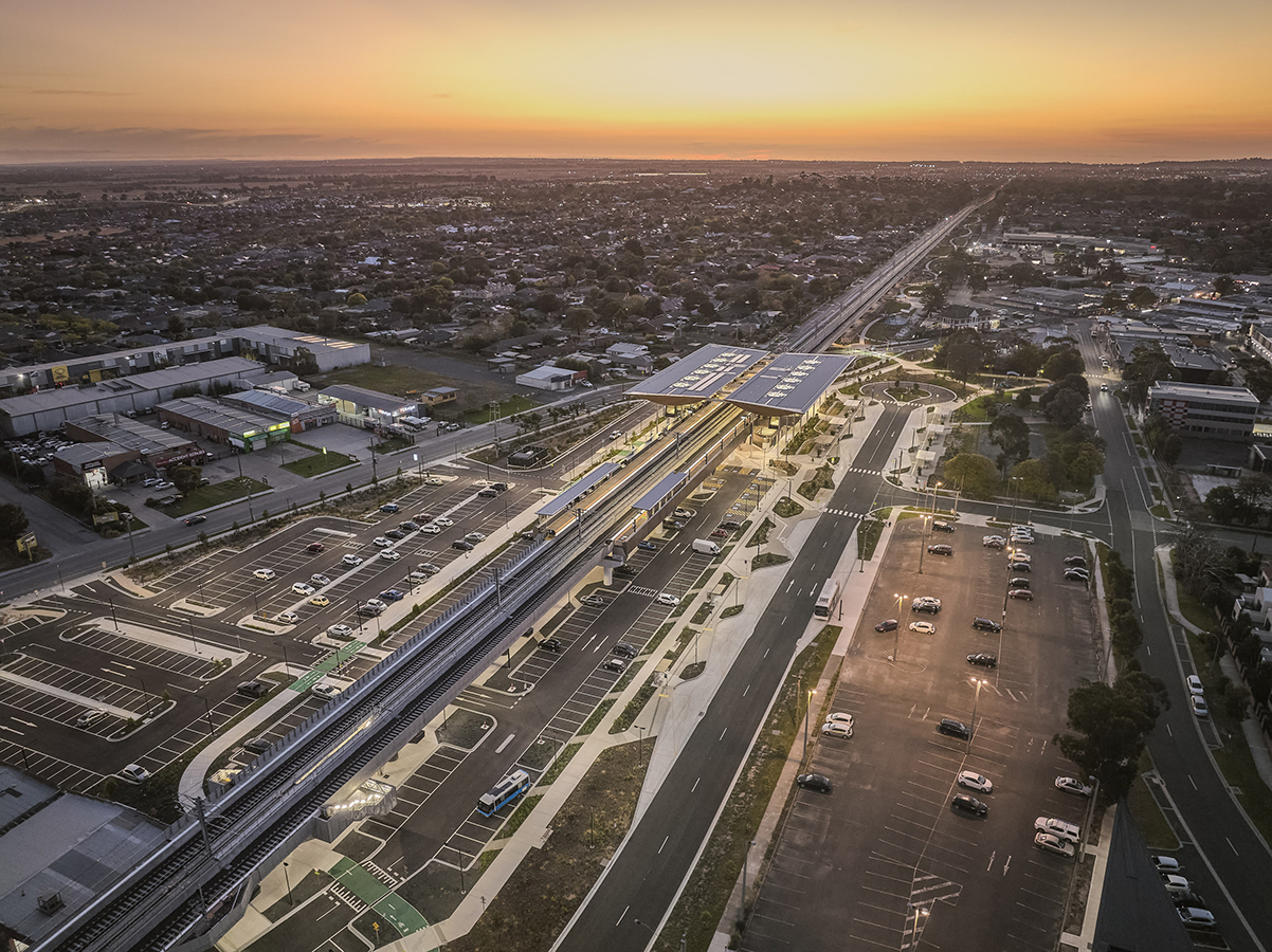 Aerial view of Pakenham Station at sunset with long rail line, car parks, urban streets, and glowing sky on horizon.