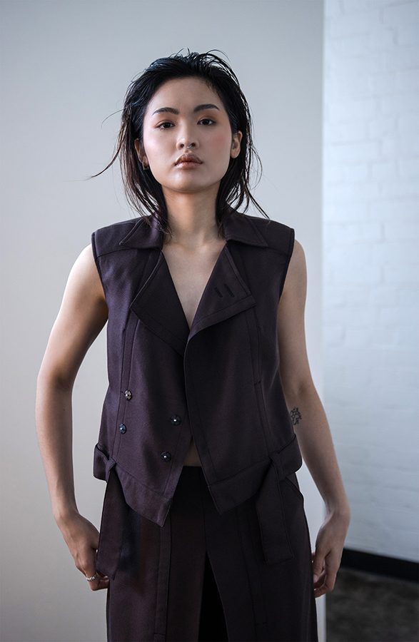 Woman in dark sleeveless vest and wide-leg trousers stands beside white geometric forms in minimalist concrete space.