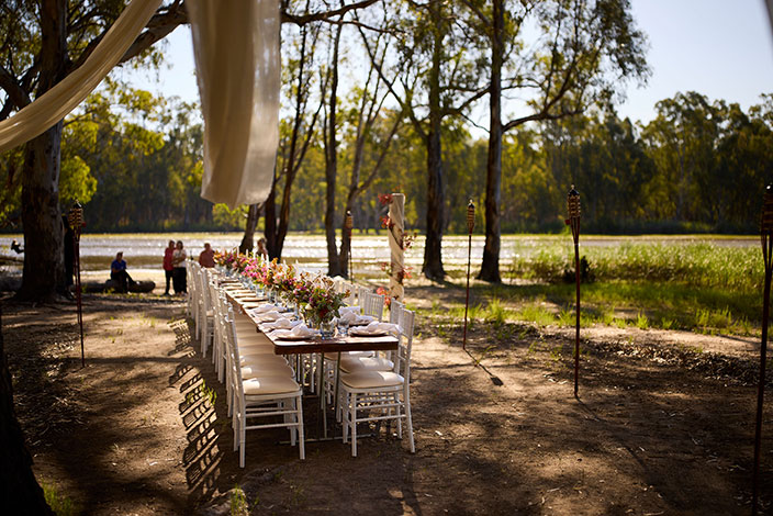 A long table with flowers is set for a community meal under river gum trees beside the water, ready to welcome guests.