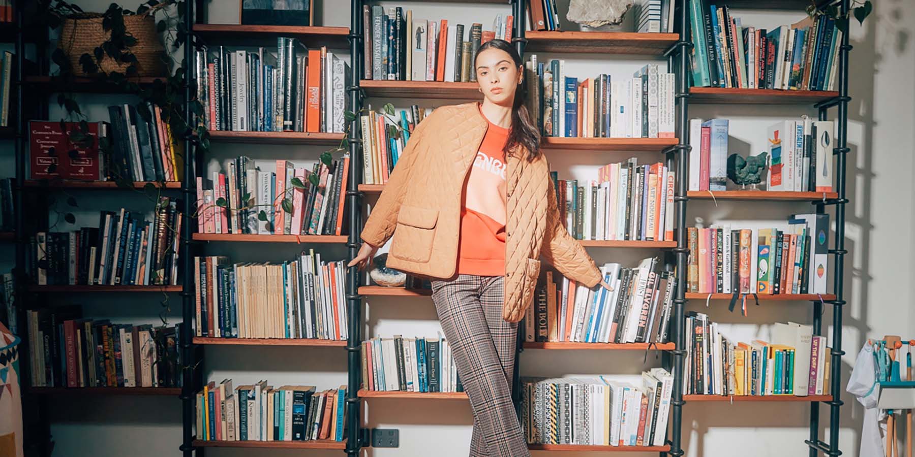 Person standing in front of a book shelf wearing a jumper, jacket and pants.