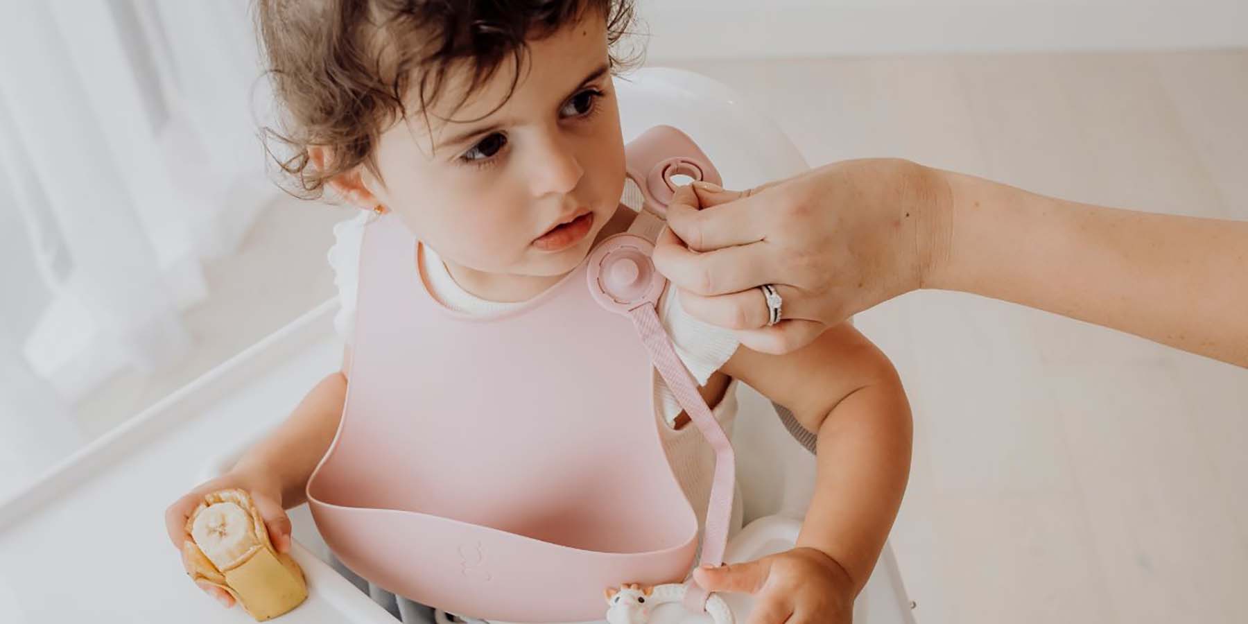 A young girl wearing a Gigi Silicone Bib, with an Accessory tether attached. A hand is gently undoing the clip with one hand.