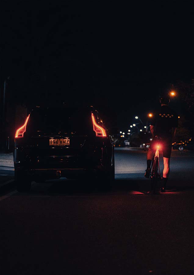 A cyclist stopped next to a car at night with a Flock Light shining light onto their legs.