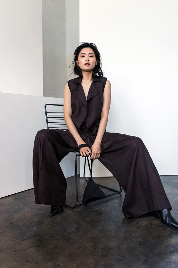 Woman in dark tailored vest and wide-leg trousers sits on black chair against minimalist white and grey concrete backdrop.