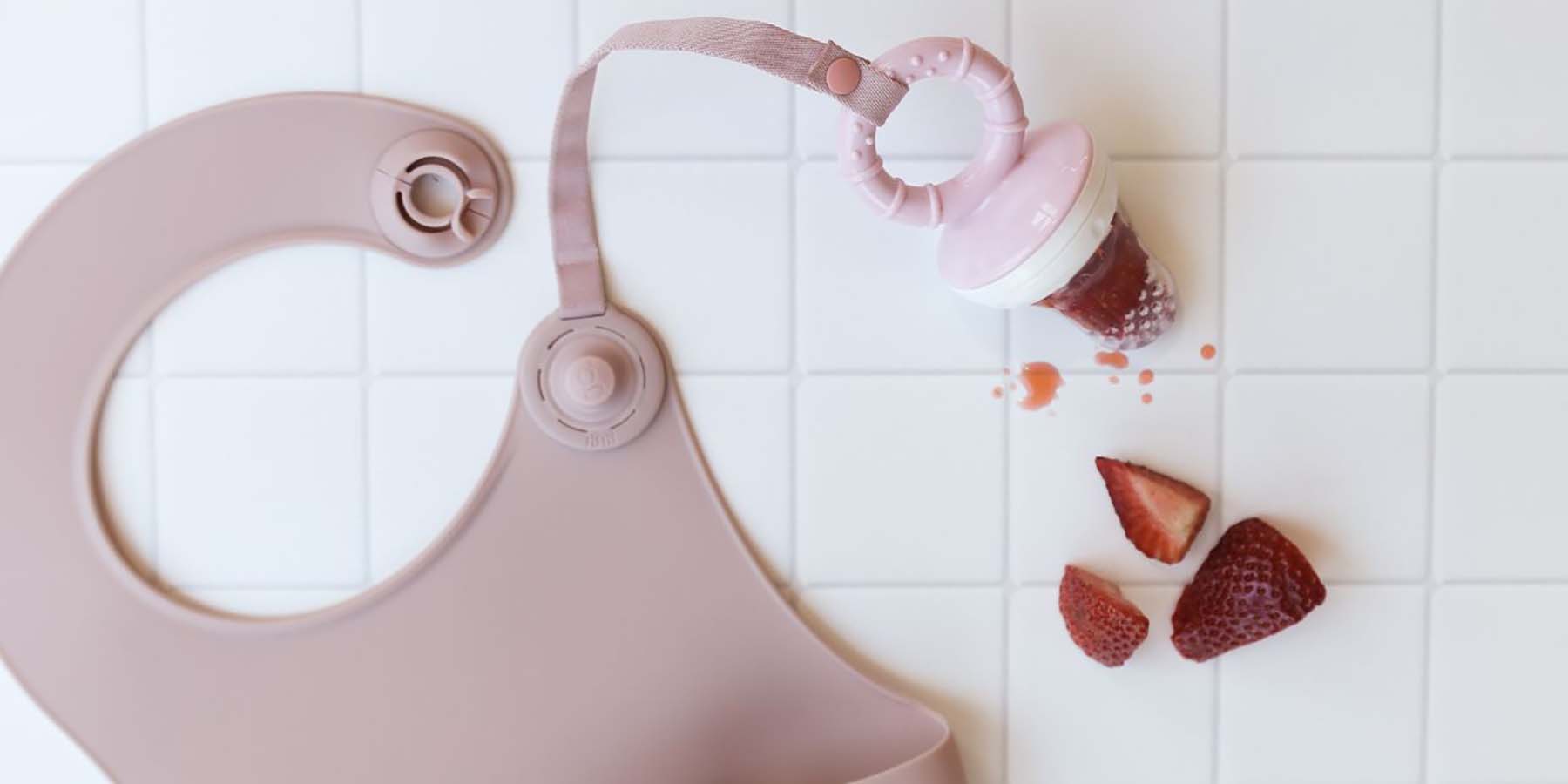 A pink silicone bib on white tiled background, with an Accessory Tether attached and strawberry inside a silicone feeder