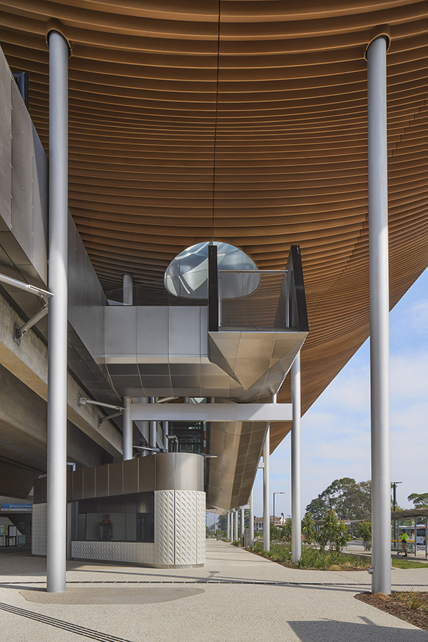 Under Pakenham Station’s timber roof with tall columns, metal details, skylight, and landscaped path to the distance.