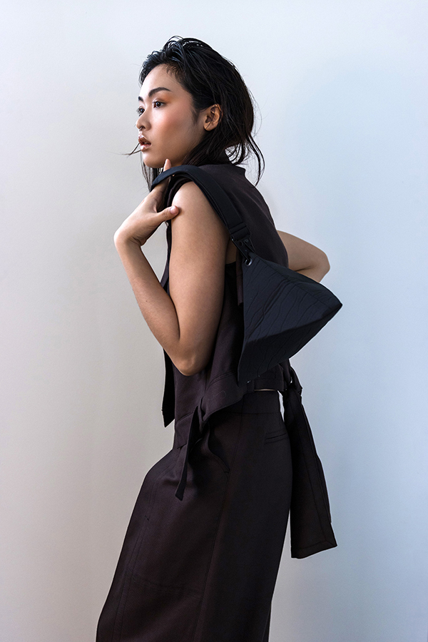 Woman in dark sleeveless vest and wide-leg trousers stands beside white geometric forms in minimalist concrete space.