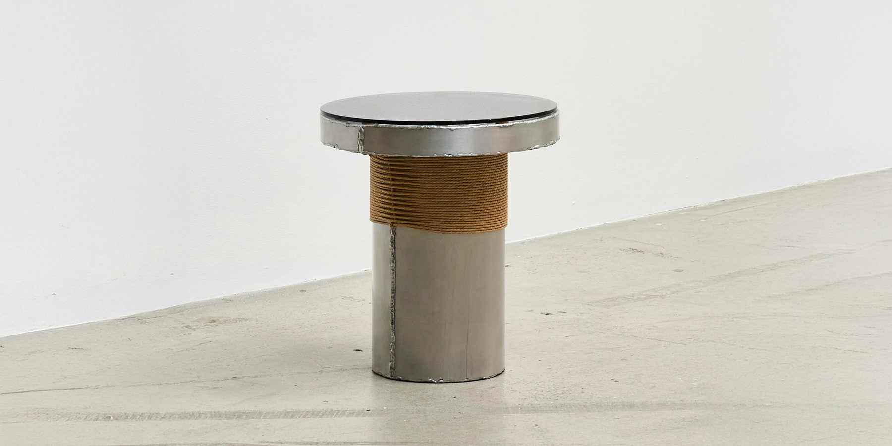 Bound Table by Locki Humphrey; a round aluminium side table with pronounced weld lines, a glass top and paper cord wrap.