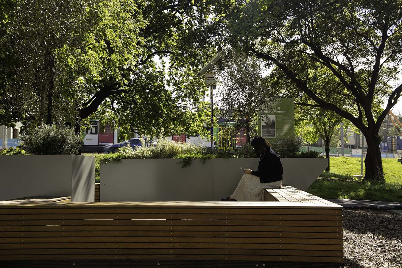 An image of a person sitting on a park bench, by planter boxes filled with plants, on a sunny day