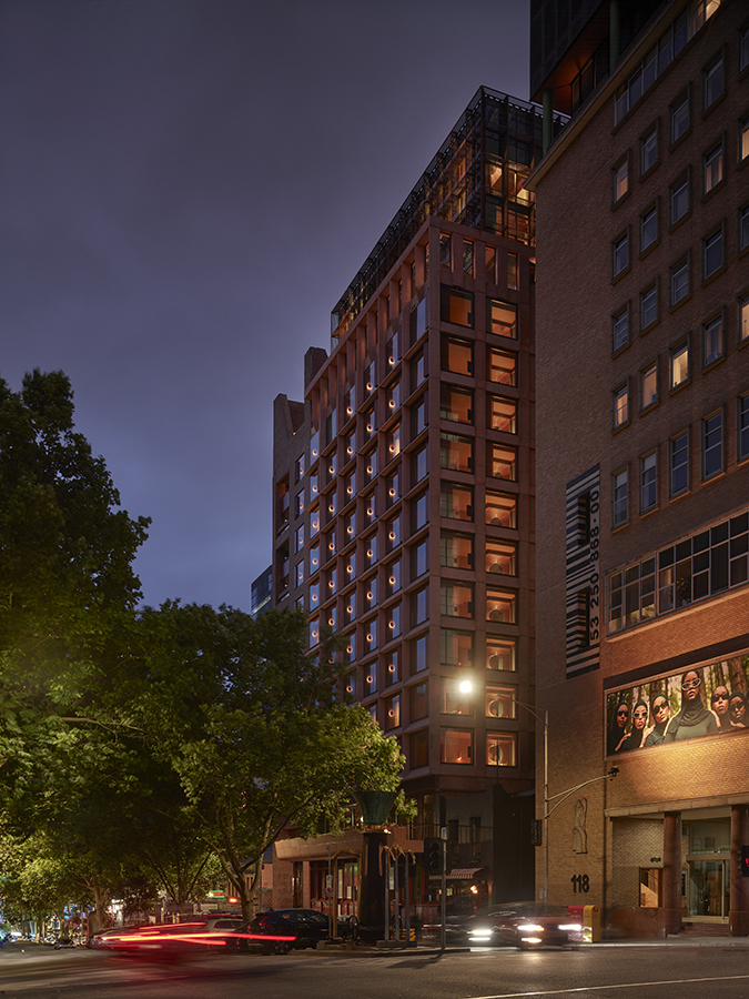 A reddish hotel building sits on a busy street lined with trees in the evening.