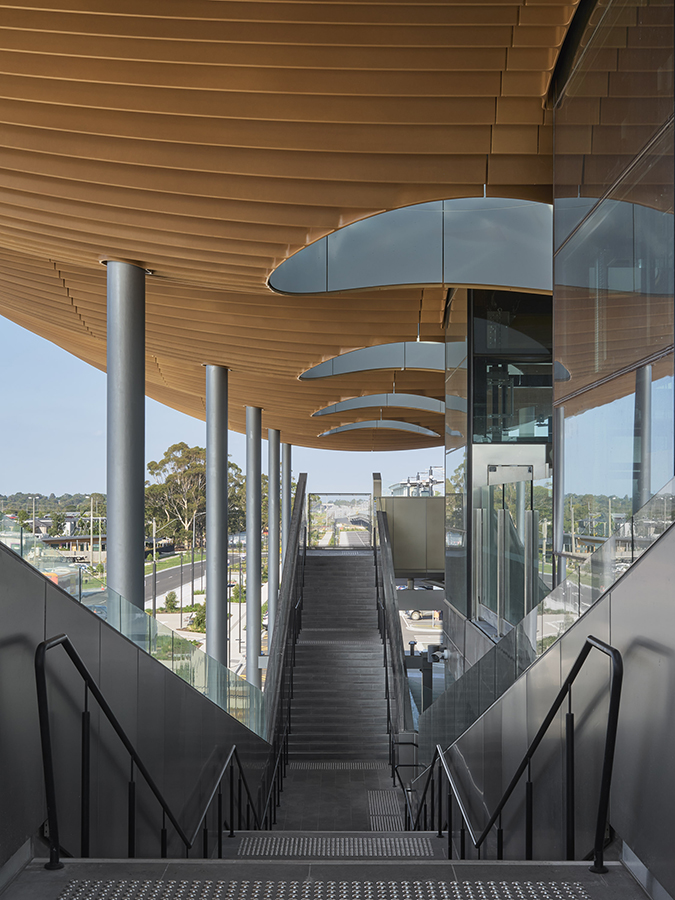 Staircase at Pakenham Station with timber-look ceiling, glass walls, sleek columns, and views to trees and streetscape.