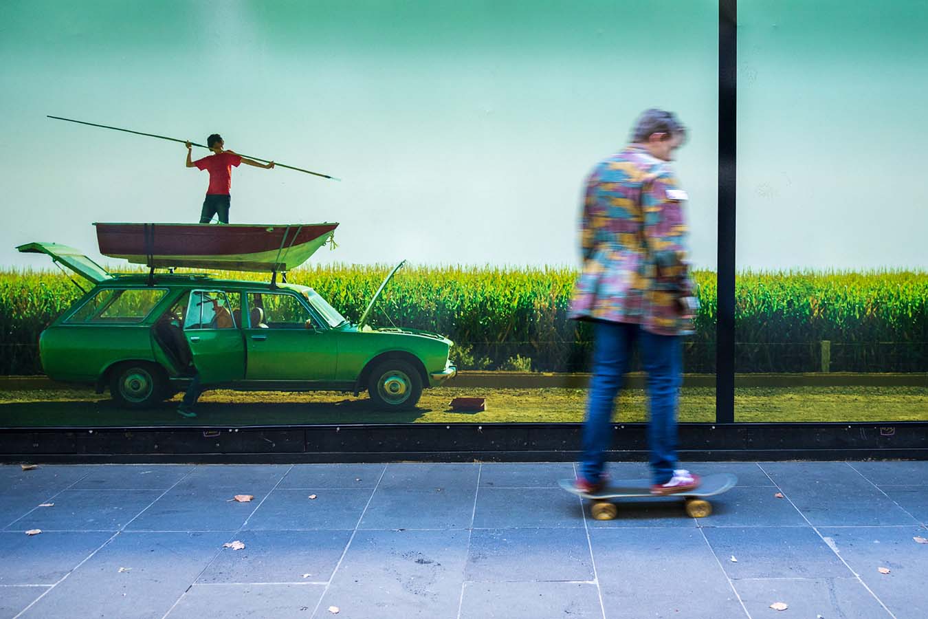 A person in a colourful suit jacket and jeans skateboards past a photograph of a boy with spear, standing in a boat on a car