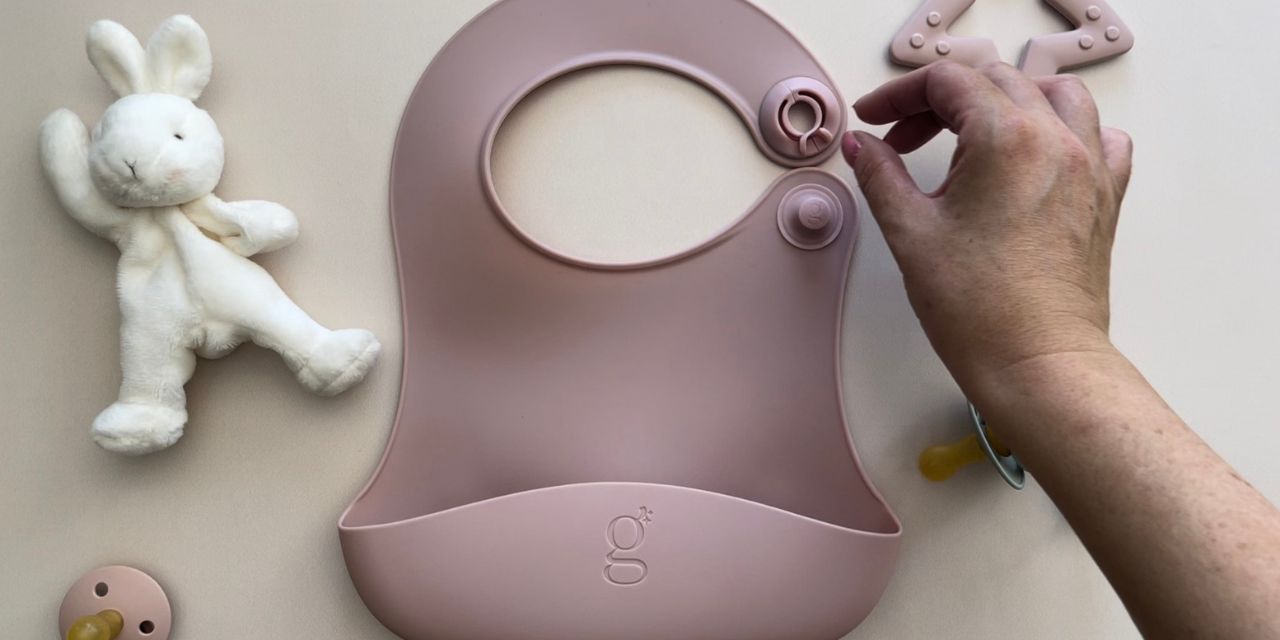 Pink Gigi Silicone Bib on a table, with a bunny comforter, dummy and teether nearby. A hand is ready to close the bib's clip