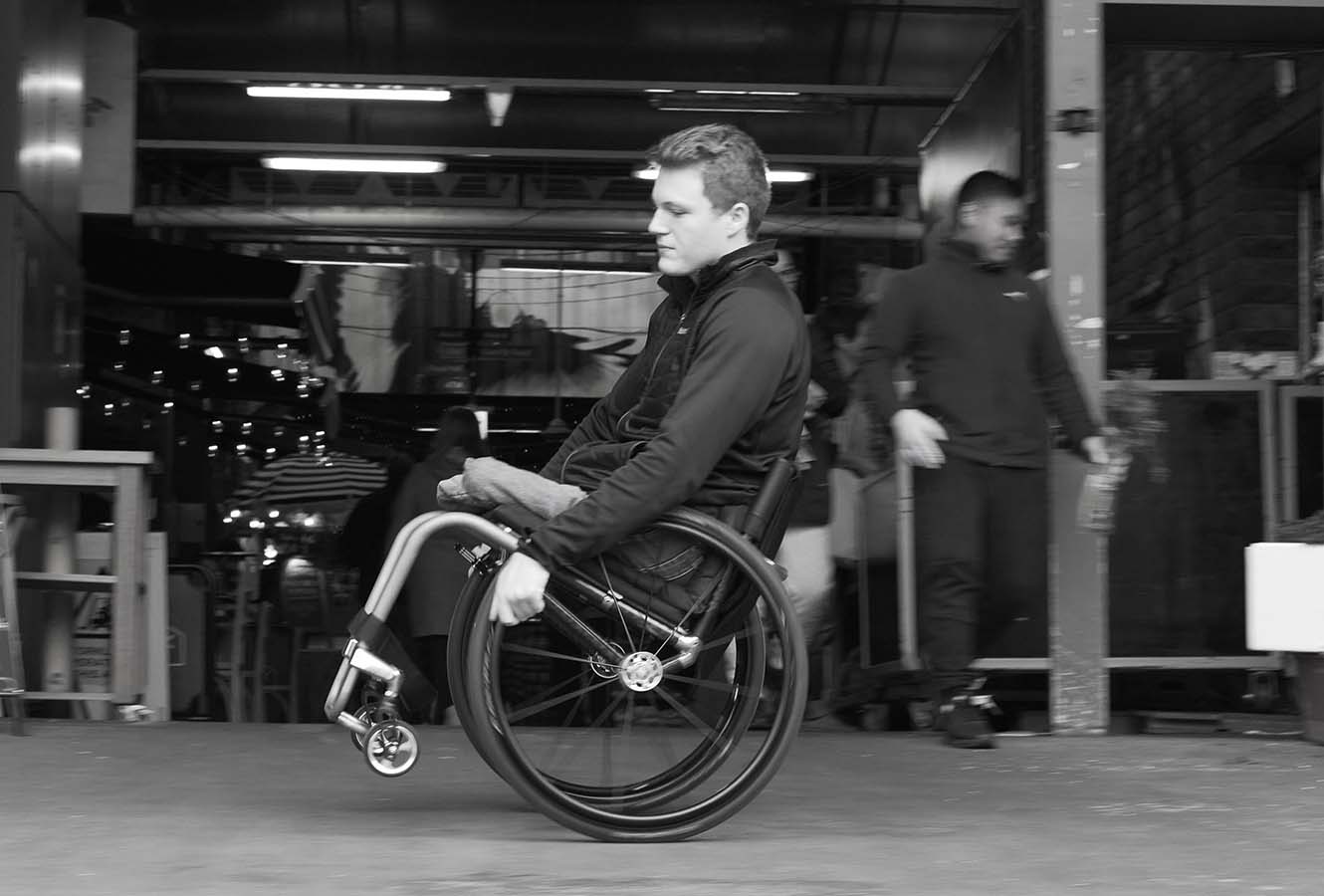 Image of a young male, doing a wheelie in a Rove Wheelchair at a market.