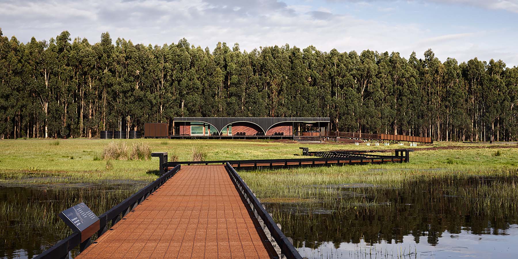 Tae Rak Aquaculture Centre and its red gum facade set behind charred timber arches, with the jetty and lake in foreground.