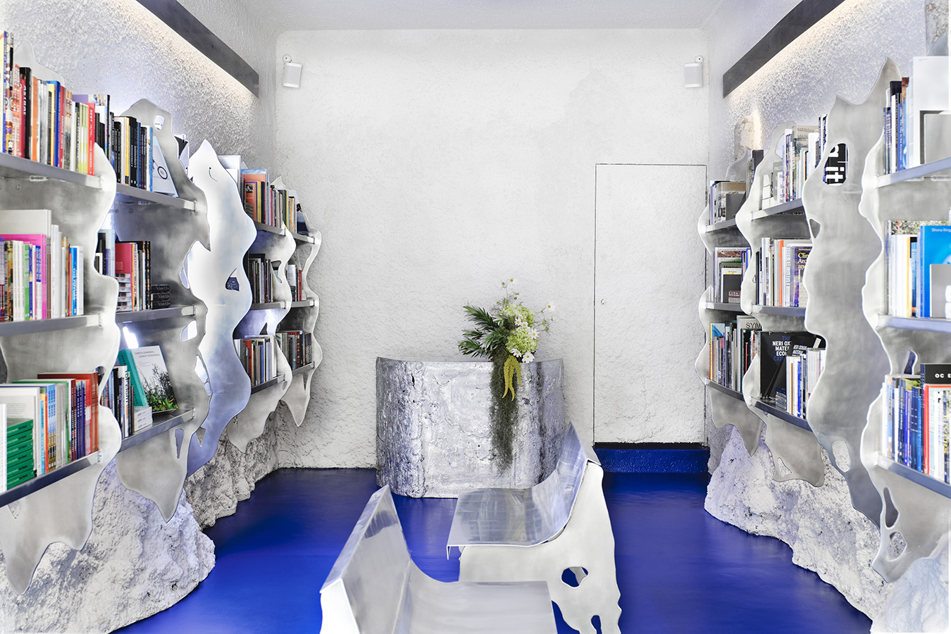 Contemporary bookshop interior with blue floor, aluminium benches, bookshelves, and shop counter with green floristry.