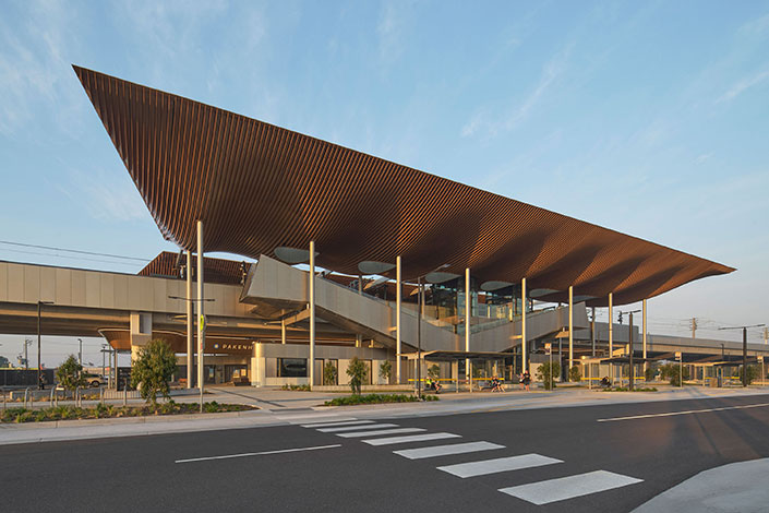 Modern train station with striking angled roof, timber-like fins, elevated platforms, and landscaping under clear sky.