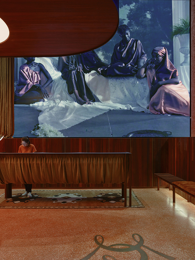 An entrance foyer, with a long tactile bench, and a large screen on the wall displays a scene of people gathering.
