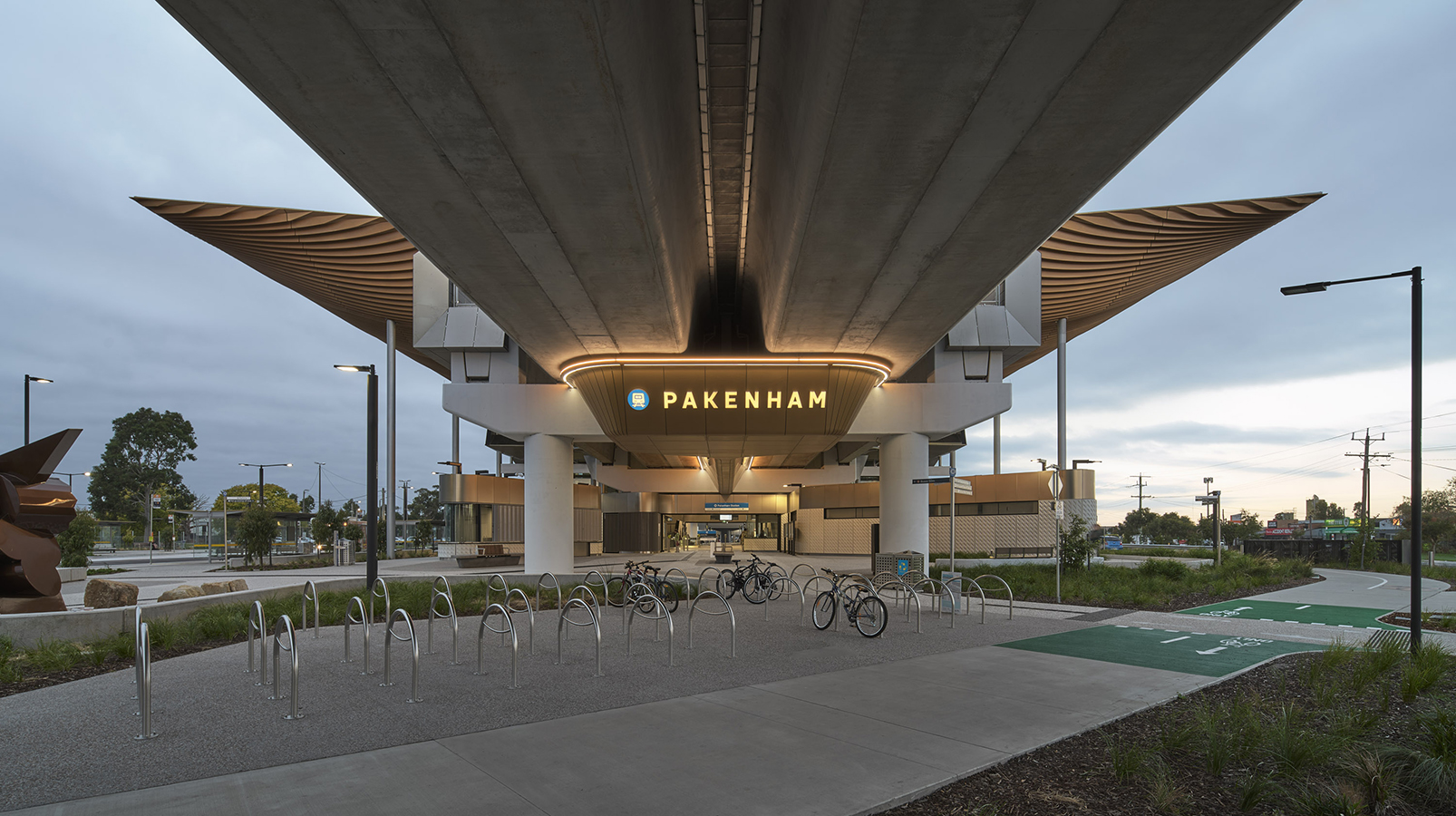 View under elevated Pakenham Station with timber roof, bike racks, lit sign, and landscaped paths at dusk.