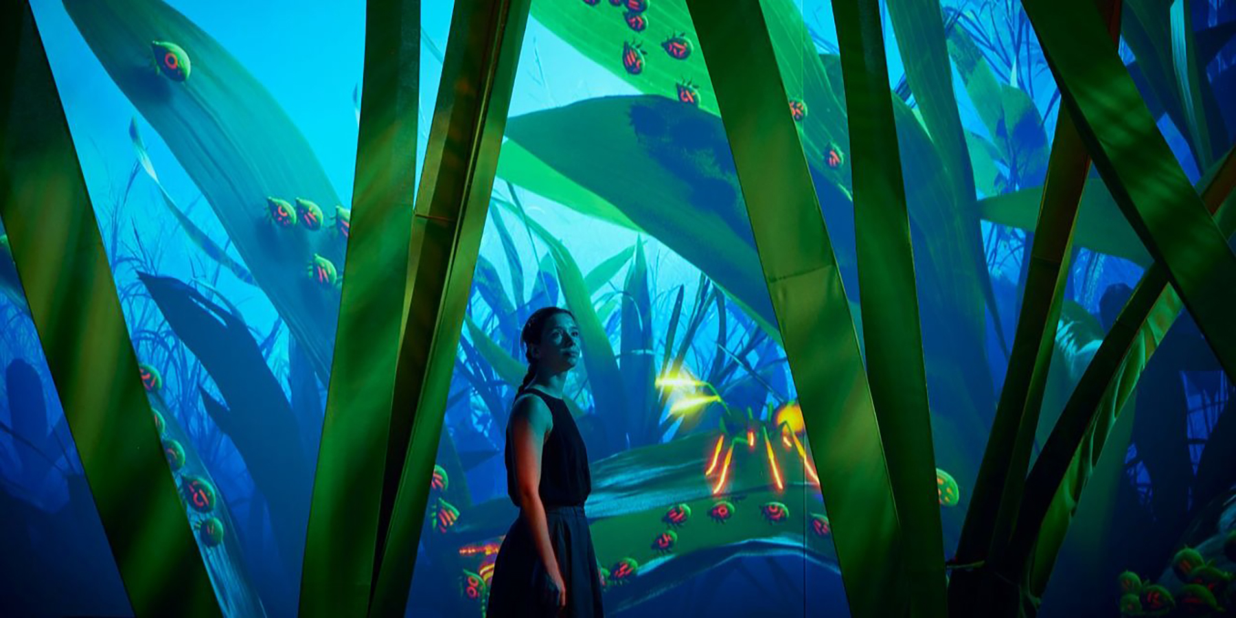 An exhibition visitor walking amongst giant blades of grass, watching ants and aphids interact