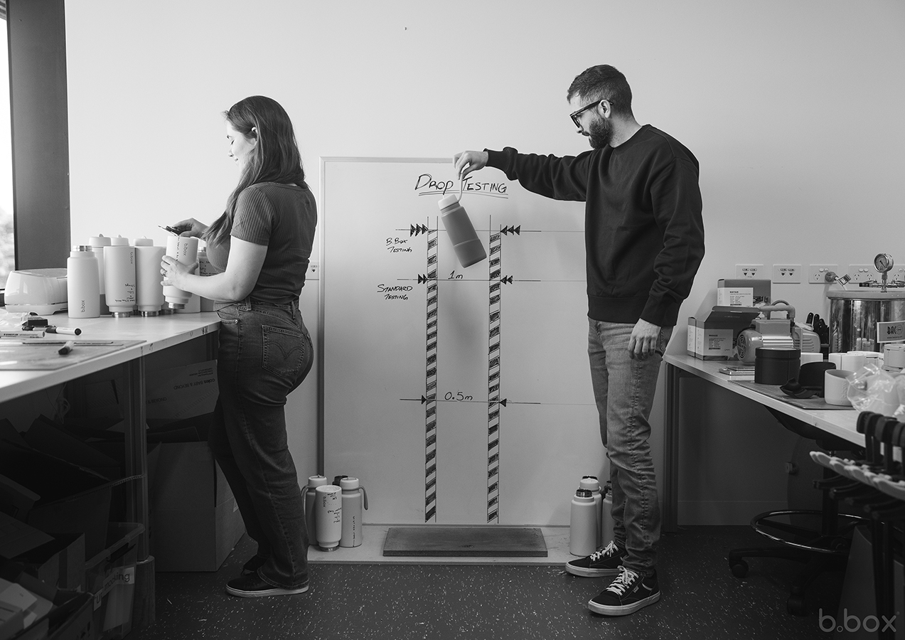 Two people drop test b.box bottles in a lab, measuring durability at 0.5m and 1m heights against a marked test board.
