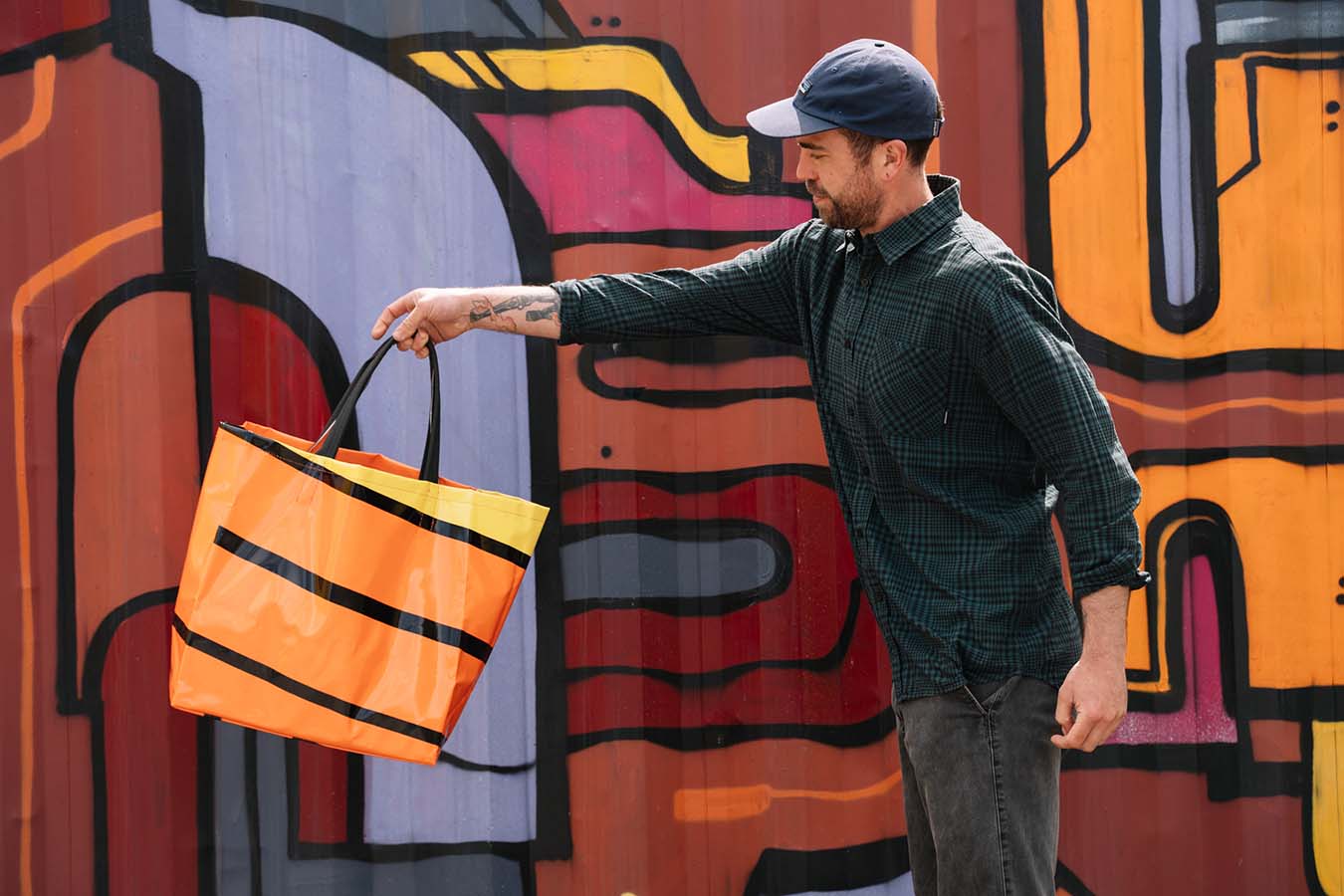 A man in a baseball cap holds up an orange and black bag. He is standing in front of a colourful artwork.