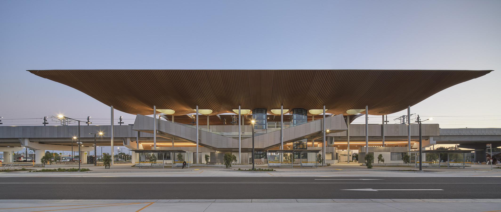Front view of Pakenham Station with large timber-lined roof, slender columns, elevated platforms, and evening lighting.