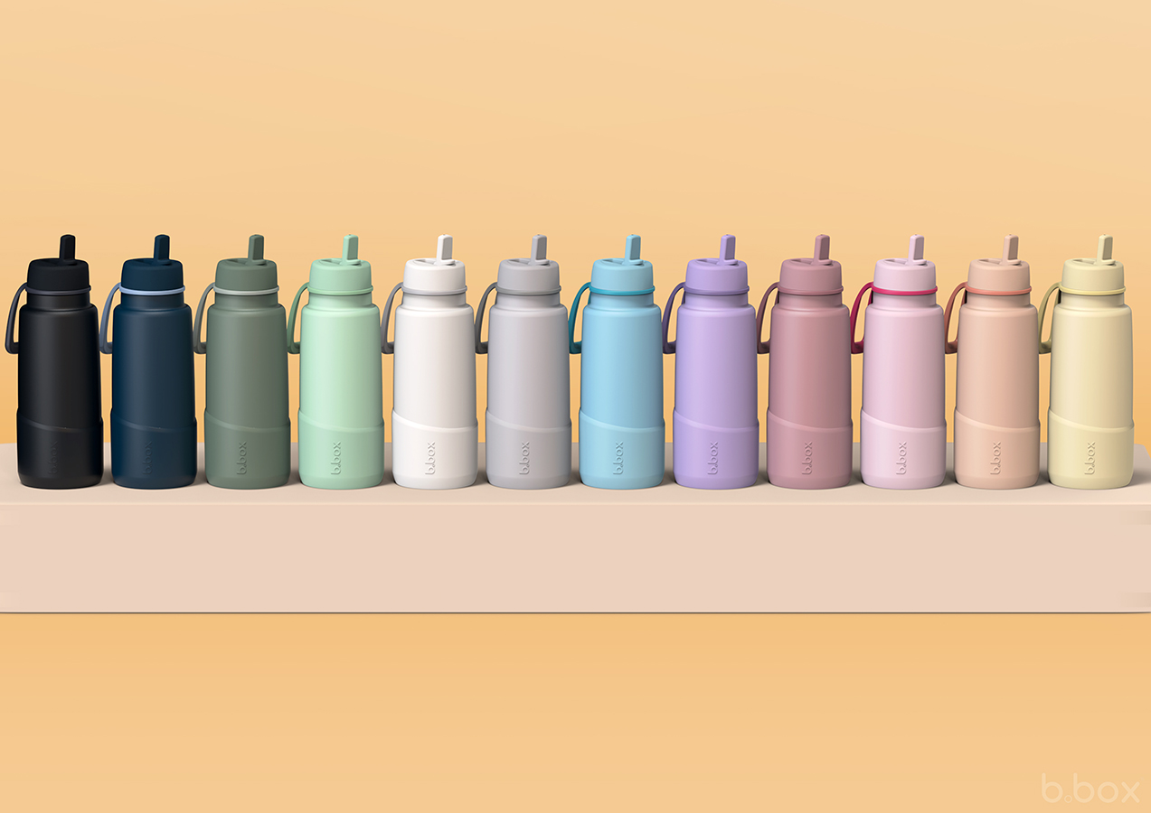 Row of b.box insulated bottles in bold colours with silicone bumper sleeves, displayed on a beige platform.