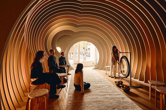People seated in a curved room while a woman plays a large gong.
