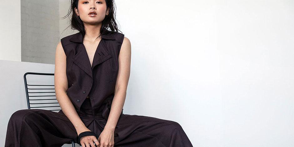 Woman in dark tailored vest and wide-leg trousers sits on black chair against minimalist white and grey concrete backdrop.