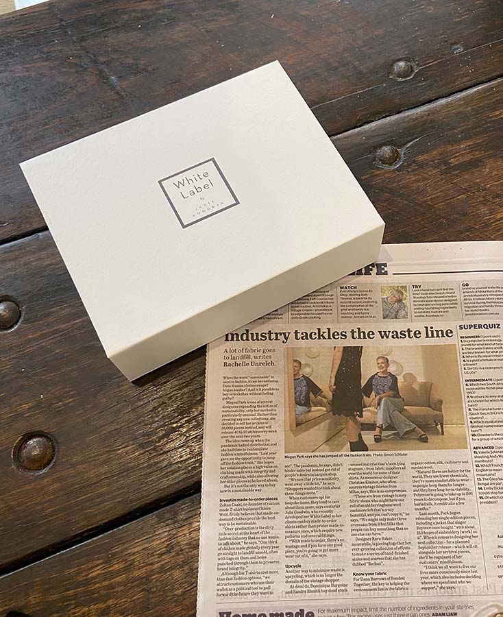 Image of The Age newspaper open on a wooden table with a White Label box beside it. Headline reads: Industry Tackles Waste.