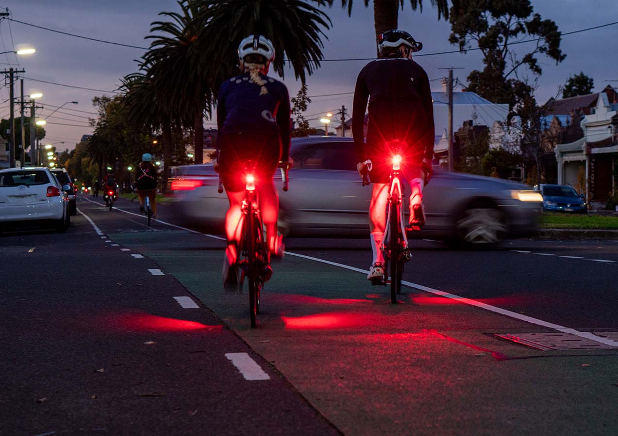 Two cyclists riding on a road at night with a Flock Light shining light onto their legs.