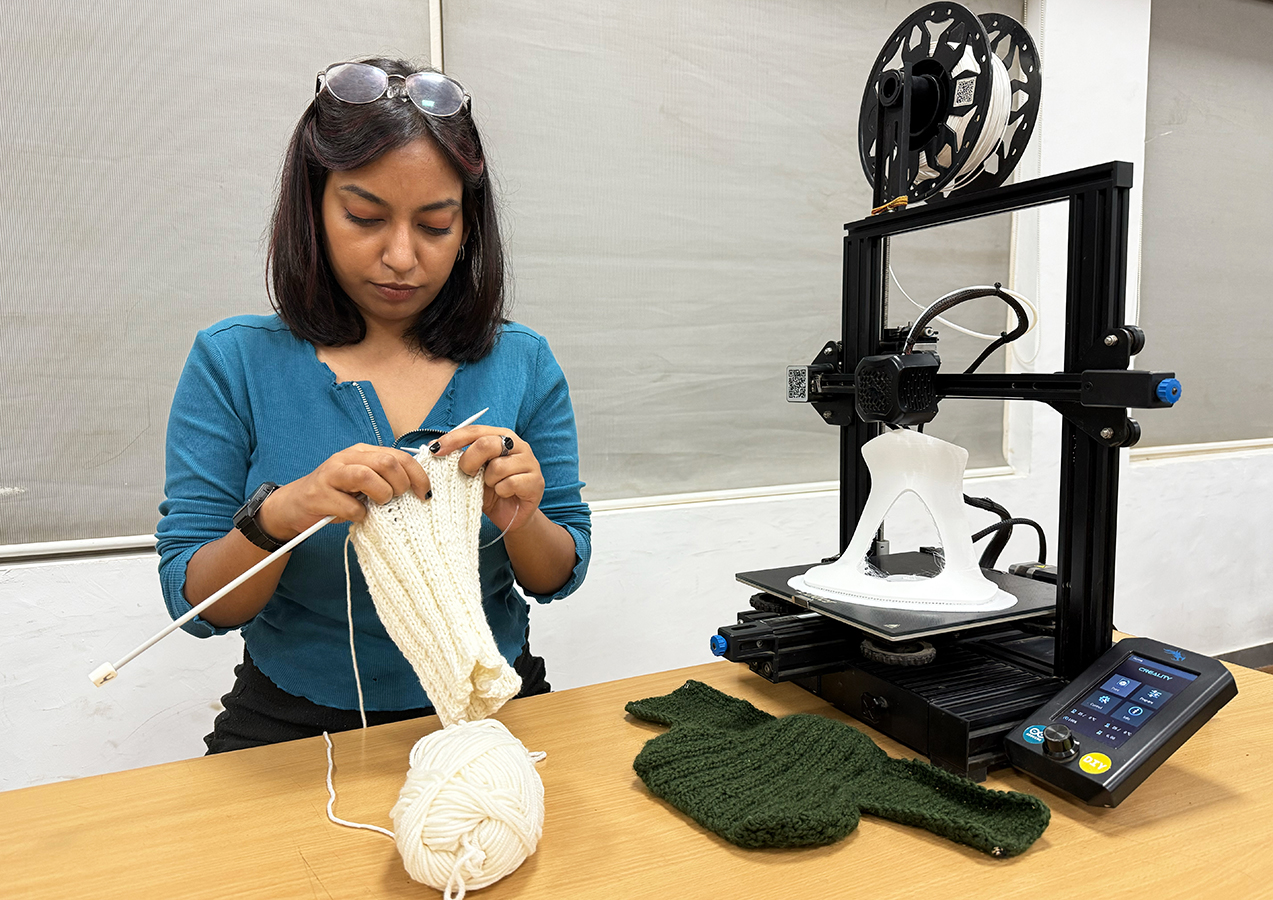 A woman knitting a white textile beside a 3D printer producing a white collar frame; green knitted parts rest on the table.