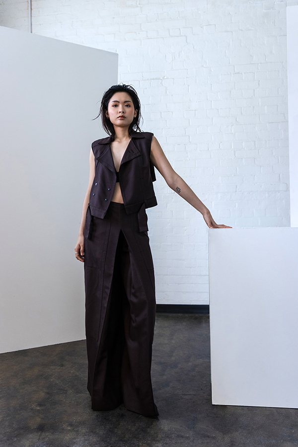 Woman in dark sleeveless vest and wide-leg trousers stands beside white geometric forms in minimalist concrete space.