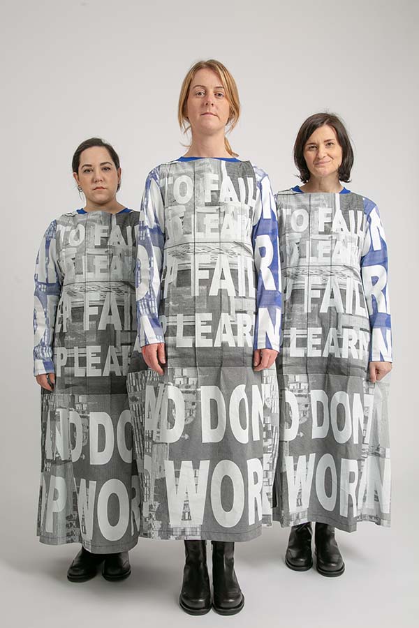Three women stand together wearing long dresses, long sleeved tops and black boots. The clothes are printed with large text