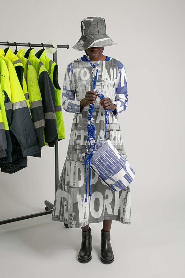A model wears a long dress, long-sleeved tee, hat and bag with printed with large letters. High-vis jackets hang beside her