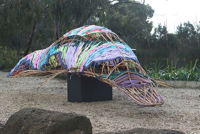 Cane and bamboos sculpture of a platypus, with pastel coloured fabric woven through the cane. Sits on a plinth next to a lake