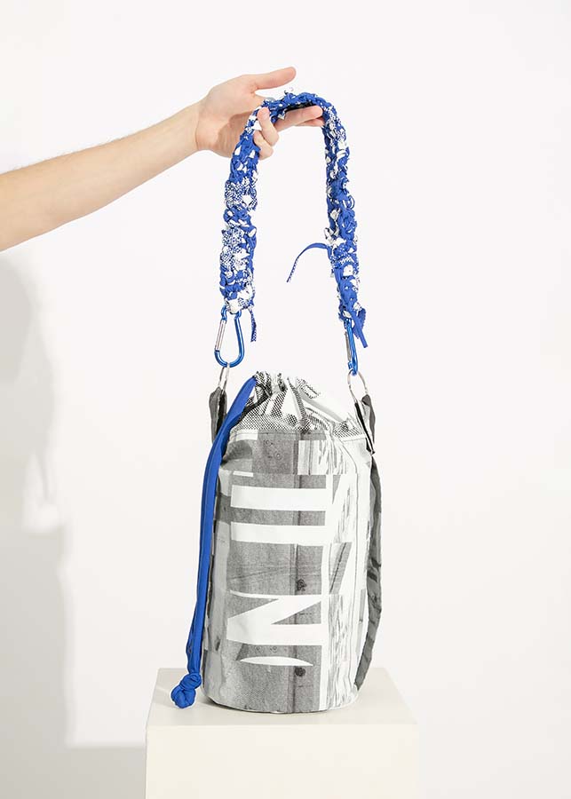 A person holds out a black drawstring bag printed with large white lettering. The strap is crocheted from a blue fabric